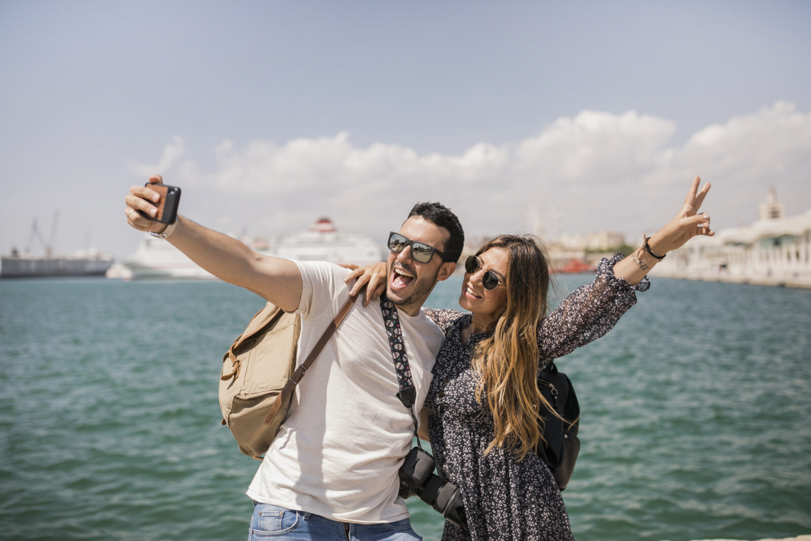 Woman making a peace sign with her boyfriend, who is taking a selfie with his cell phone.